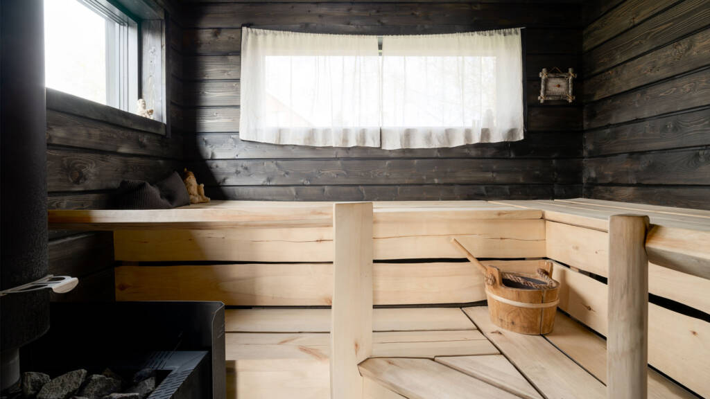 Tumma sauna, jossa vaaleat lauteet ja kaksi ikkunaa valkoisin verhoin.