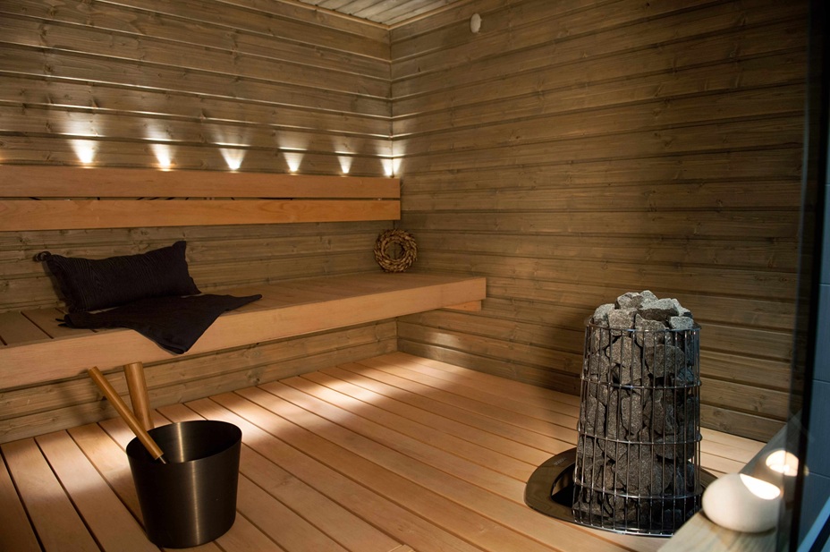 Tumma kuultokäsitelty sauna – vaaleat lauteet, häkkikiuas ja LED-spotit.