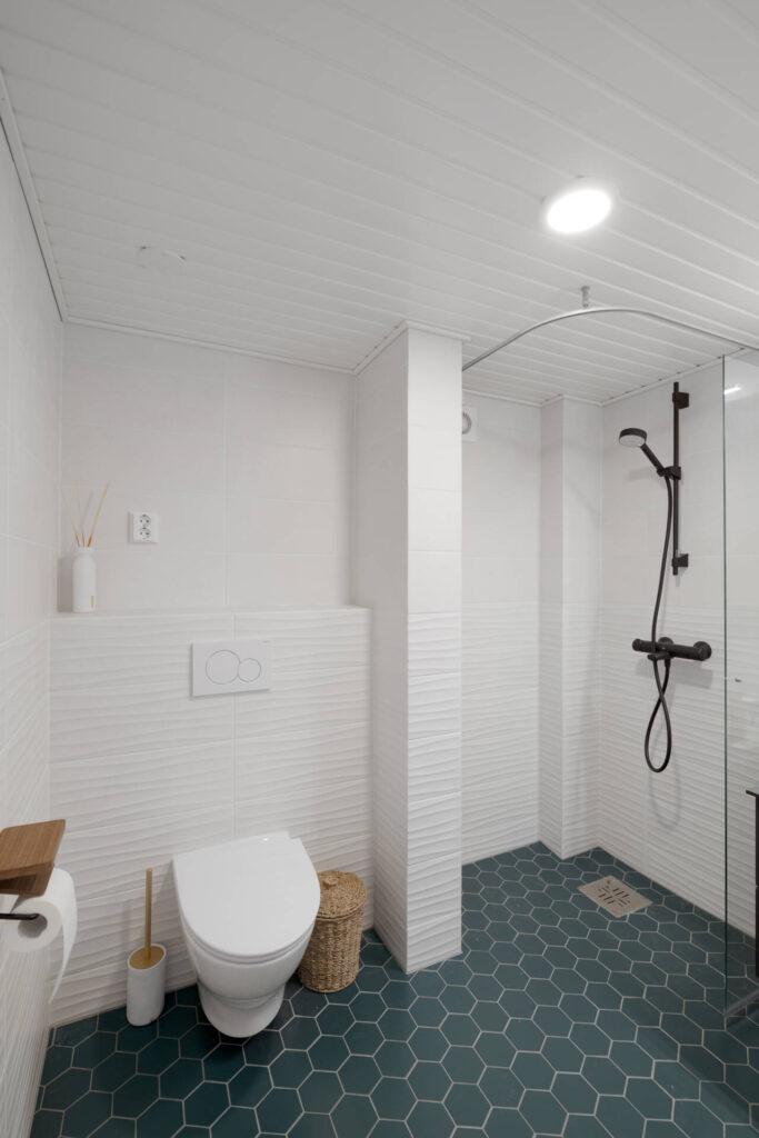 Valkoinen WC, jossa aaltokuvioinen seinälaatta, tumma kuusikulmiolattia ja musta suihku.